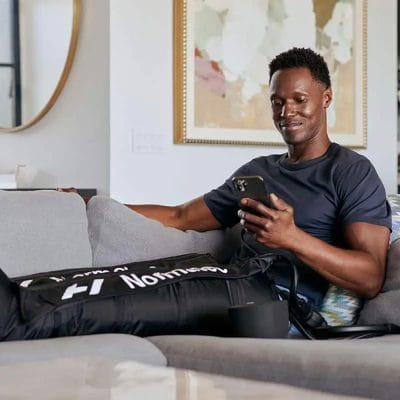 Male athlete sitting on sofa using Hyperice Normatec 3 Legs compression boots for leg recovery and performance